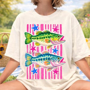 May include: White t-shirt featuring two colorful fish illustrations. The fish are green and blue, with pink mouths and yellow accents. The design includes pink vertical stripes and blue and pink stars.