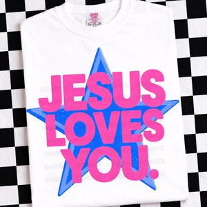 May include: White t-shirt with a blue star and the text "JESUS LOVES YOU" in pink. The shirt has a crew neck and short sleeves. The shirt is made of a soft, comfortable material.