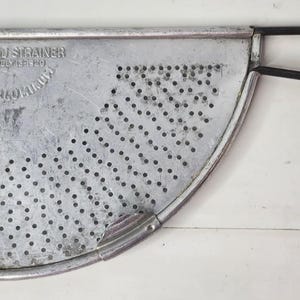 以下が含まれることがあります： 長い黒いワイヤーハンドルが付いた半円形のアルミニウム製ポットストレーナー。金属面には穴あき模様があり、FOLEY POT STRAINER, MADE IN U.S.A.という文字が刻印されています。このキッチンツールには、湾曲した端に2つの小さな金属製クリップが付いています。