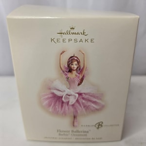 May include: A Hallmark Keepsake Flower Ballerina Barbie ornament box featuring a graphic of a doll in a pink tutu with white tulle accents. The cream-colored box displays the Hallmark logo, the text Flower Ballerina Barbie Ornament, and a Barbie B logo.