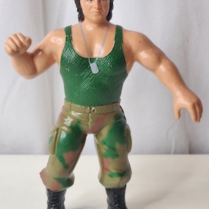 Könnte beinhalten: Eine Kunststoff-Actionfigur eines Soldaten mit bordeauxrotem Barett, grünem, texturiertem Tanktop und Camouflage-Hose. Die Figur hat dunkles Haar, eine silberne Erkennungsmarke an einer Kette und schwarze Stiefel, ein Arm ist erhoben.