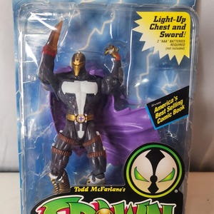 Puede incluir: Una figura de acción Redeemer en su empaque original. La figura presenta un traje negro y dorado, una capa morada y una máscara dorada. El empaque incluye textos como Redeemer, Spawn, Deluxe Edition y Light-Up Chest and Sword.