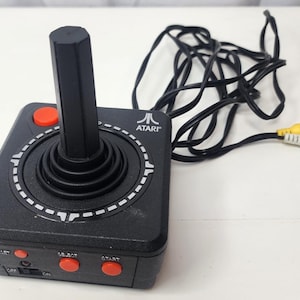 May include: A black Atari joystick controller featuring a central vertical stick, a red button on the top surface, and three smaller red buttons labeled Reset, Select, and Start on the front edge. It includes a black cord with yellow and white RCA plugs.