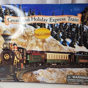 Puede incluir: Una caja de cartón rectangular para un set de tren GreatLand Holiday Express de 1996. La caja presenta un gráfico de una locomotora de vapor negra, un vagón de pasajeros crema y verde y un furgón de cola verde sobre vías en la montaña.