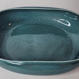 May include: An oval-shaped ceramic bowl with a deep teal, glossy glaze. The bowl features a smooth, continuous rim and a wide, open basin. The interior surface shows subtle variations in the blue-green color, reflecting light across the curved walls.