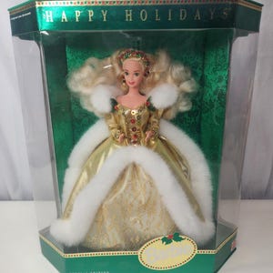 Pode incluir: Uma boneca loira num vestido dourado com acabamento em pele sintética branca, lantejoulas vermelhas e verdes e um toucado a condizer, embalada numa caixa verde com a etiqueta Happy Holidays. A boneca tem cabelo comprido e ondulado.