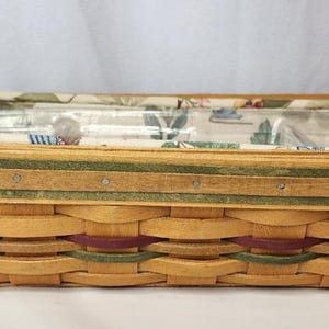 Puede incluir: Una cesta tejida rectangular y alargada con tiras de madera de color tostado claro, acentuada con bandas horizontales de color verde oscuro y rojo intenso. La cesta incluye dos asas curvas y un forro de tela con estampado floral.