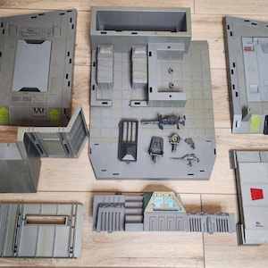 Pode incluir: Um conjunto de componentes de brinquedo em plástico cinzento, incluindo painéis de parede, uma base com estruturas de salas, uma plataforma de aterragem com marcas vermelhas e um símbolo H, e vários pequenos acessórios cinzentos.