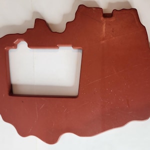 Puede incluir: Un componente plano de plástico de color marrón rojizo con una forma exterior orgánica e irregular y un gran recorte rectangular en el centro. La superficie presenta ligeros arañazos y pequeñas pestañas que sobresalen en los bordes.
