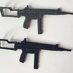 May include: Two miniature toy rifles, one in dark gray and one in black. Each plastic toy features a long barrel, a textured pistol grip, a vertical magazine, and a skeletonized stock. The rifles are identical in design and scale, presented side by side.
