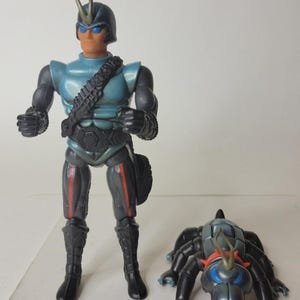 May include: A standing action figure with a blue helmet, blue chest armor, and black limbs, accompanied by a smaller, multi-legged black and blue creature figure. The humanoid figure features a tan face, blue eyes, and a prominent antenna on the helmet.