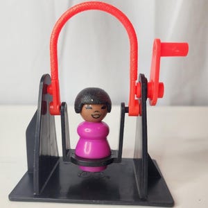 May include: A plastic toy swing set featuring a black base and frame with a bright red arch and hand crank. A small, stylized figure with a dark brown complexion, black hair, and a pink torso is seated in the center of the black swing seat.