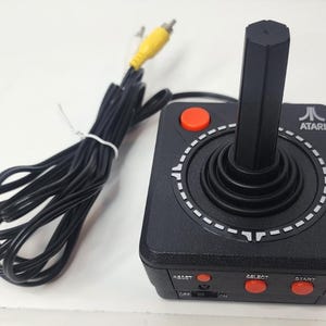 Puede incluir: Un mando de joystick Atari negro con una palanca vertical alta, un botón rojo en la parte superior y tres botones rojos más pequeños etiquetados como Reset, Select y Start en la parte frontal. Incluye un cable negro largo con un conector RCA amarillo.