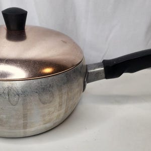 May include: A round aluminium saucepan with a polished copper lid featuring a black knob. The pan has a black handle with a small metal hanging loop at the end. The exterior of the aluminium base shows signs of wear and surface scratches.