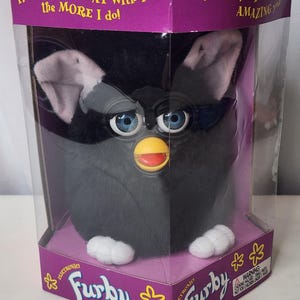 May include: A black Furby electronic toy with blue eyes, pink-lined ears, and white feet, packaged in a purple box. The box features the text: The more you PLAY with me the MORE I do! I will keep AMAZING you! and the Furby logo with yellow flowers.