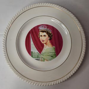 Op de afbeelding: Een ronde witte keramische herdenkingsbord met een centraal rond portret van koningin Elizabeth II, gekleed in een kroon en een lichtgroene jurk tegen een rode gordijnachtergrond. Het bord heeft een gedraaide rand en een gouden bies.
