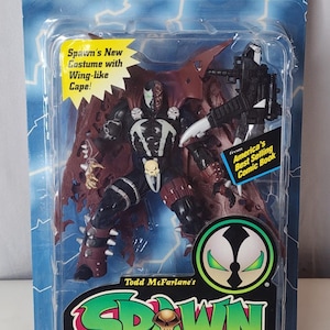 Puede incluir: Una figura de acción Spawn II en su empaque blíster original. La figura presenta un traje negro y granate con una capa en forma de alas, detalles de calaveras y un accesorio de arma. El empaque incluye el texto Spawn II, Todd McFarlane's y Deluxe Edition.