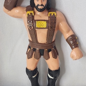 Può includere: Un action figure in plastica di un wrestler con capelli scuri e barba, che indossa cinghie in similpelle marrone con fibbie gialle, una gonna marrone, slip neri, polsini neri e stivali neri stringati. Il personaggio ha un braccio alzato.
