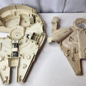 Può includere: Un modello di astronave in plastica beige smontato, caratterizzato da uno scafo principale circolare con scomparti interni e una sezione superiore con dettagli stampati, incluso un componente della cabina blu scuro sul lato sinistro.
