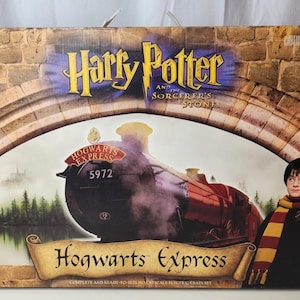 Puede incluir: Una caja de cartón rectangular para un set de tren Hogwarts Express de Harry Potter y la piedra filosofal. La caja muestra una locomotora de vapor roja numerada 5972, una ilustración de Harry Potter con una lechuza blanca y arcos de piedra.