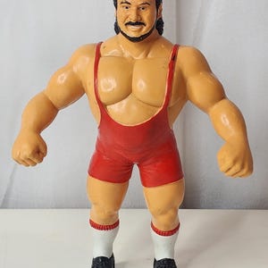 以下が含まれることがあります： 黒い髪と髭を生やした筋肉質なプロレスラーのプラスチック製アクションフィギュア。赤いシングレット、赤い縁取りのある白い靴下、黒い靴を着用している。レスラーは日焼けした肌の色をしており、直立したポーズをとっている。