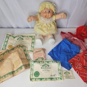 May include: A Cabbage Patch Kids doll with yellow yarn hair, wearing a yellow dress with white lace trim and white shoes. Included are a red cowboy hat, blue shorts, a red and white checkered shirt, a red bandana, and birth certificate papers.