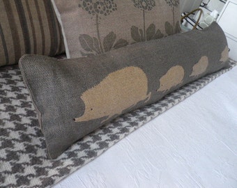 May include: A long, gray, rectangular pillow with a brown and white houndstooth pattern on one side. The other side of the pillow has a brown background with a pattern of beige hedgehogs.