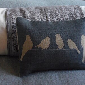 Op de afbeelding: Een rechthoekig, donkergrijs sierkussen met een beige vogelontwerp. De vogels zitten op een horizontale lijn. Het kussen ligt op een bed met andere kussens en een deken.