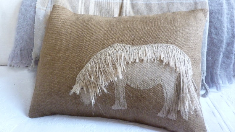 May include: A brown burlap pillow with a white horse design. The horse is made of fringe and has a long mane.