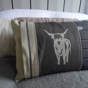May include: A decorative pillow with a gray background and a white highland cow design. The pillow has a brown, white, and gray plaid fabric on one side and a solid gray fabric on the other side.