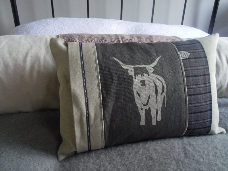 May include: A decorative pillow with a gray background and a white highland cow design. The pillow has a brown, white, and gray plaid fabric on one side and a solid gray fabric on the other side.