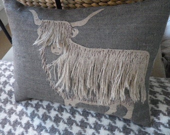 May include: A gray throw pillow with a brown and white embroidered design of a Highland cow with long hair. The pillow is on a gray and white checkered fabric.