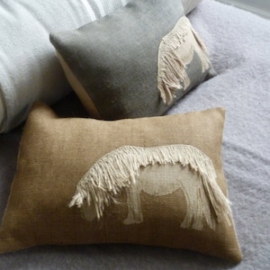 May include: Two decorative throw pillows with embroidered horse designs. The pillows are on a light gray bedspread. One pillow is a light gray linen with a white horse design. The other pillow is a brown burlap with a white horse design.