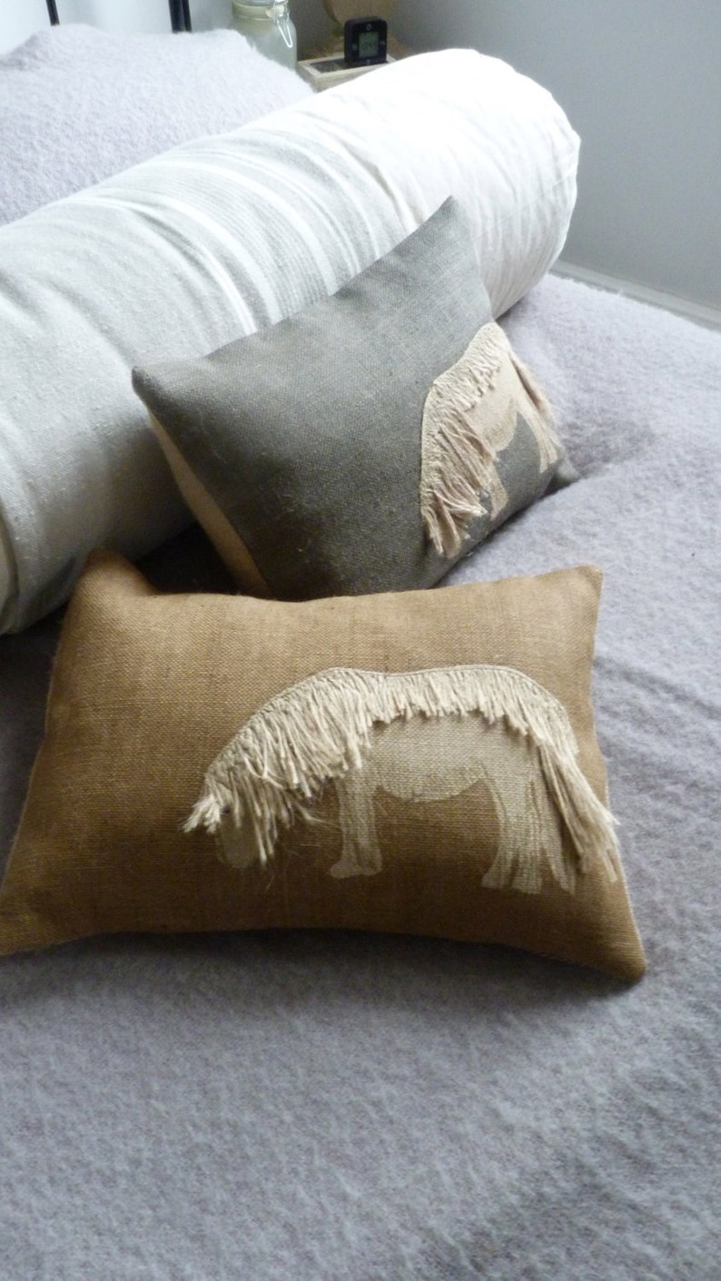 May include: Two decorative throw pillows with embroidered horse designs. The pillows are on a light gray bedspread. One pillow is a light gray linen with a white horse design. The other pillow is a brown burlap with a white horse design.