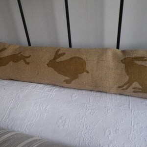 May include: A long, rectangular brown linen pillow with three brown silhouettes of rabbits printed on it. The pillow is lying on a white bedspread.