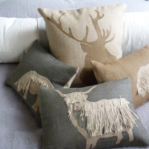 May include: Four decorative throw pillows with embroidered animal silhouettes. The pillows are made of a natural linen fabric and feature a stag, a highland cow, and two horses. The pillows are arranged on a light gray bedspread.