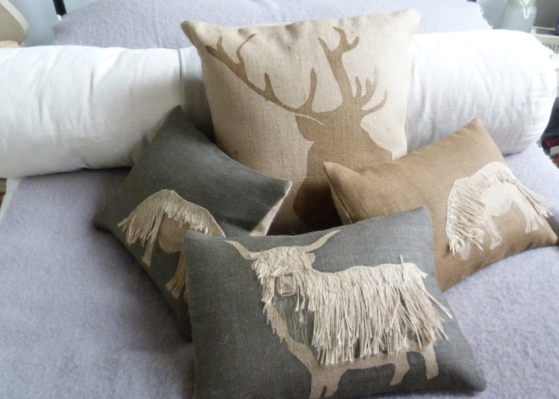 May include: Four decorative throw pillows with embroidered animal silhouettes. The pillows are made of a natural linen fabric and feature a stag, a highland cow, and two horses. The pillows are arranged on a light gray bedspread.