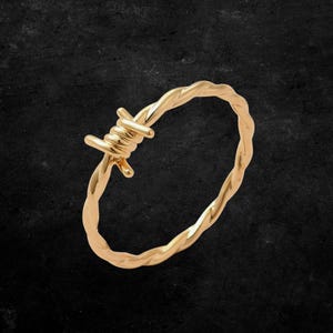 Könnte beinhalten: Ein goldfarbener Ring mit einem Stacheldraht-Design. Der Ring besteht aus einem glänzenden, metallischen Material und ist vor einem dunklen Hintergrund platziert. Das Design des Rings zeigt gedrehten Draht und scharfe Widerhaken.