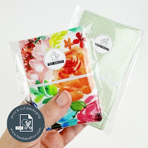 Puede incluir: Dos carteras de tela en embalaje transparente. Una cartera presenta un estampado floral con flores rosas, naranjas y azules. La otra es de color verde claro con lunares blancos. Cada cartera está asegurada con una banda elástica blanca y tiene una pegatina que dice "Tea Wallet" y "Drink Wallet".