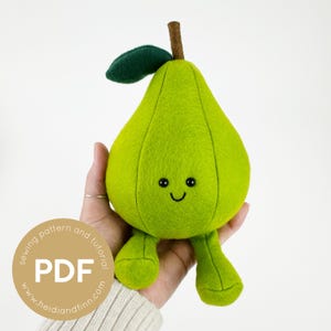 Op de afbeelding: Een groen vilten peren knuffel met een glimlachend gezicht en kleine pootjes. De peer wordt in een hand gehouden. De afbeelding bevat de tekst "sewing pattern and tutorial PDF www.heidlandfinn.com".