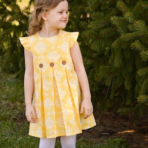 May include: A young girl wearing a yellow and white patterned dress with a ruffled neckline and three wooden buttons. The dress has pleats in the skirt and the girl is wearing white tights and brown boots.