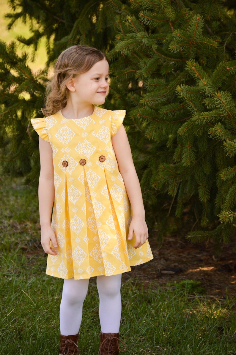 May include: A young girl wearing a yellow and white patterned dress with a ruffled neckline and three wooden buttons. The dress has pleats in the skirt and the girl is wearing white tights and brown boots.