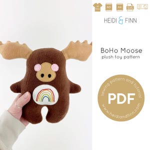 Op de afbeelding: Een bruin pluche eland speelgoed met een regenboog op zijn borst. Het speelgoed wordt vastgehouden door een hand. De afbeelding bevat ook tekst die zegt "BoHo Moose plush toy pattern sewing pattern and tutorial PDF www.heidiandfinn.com"