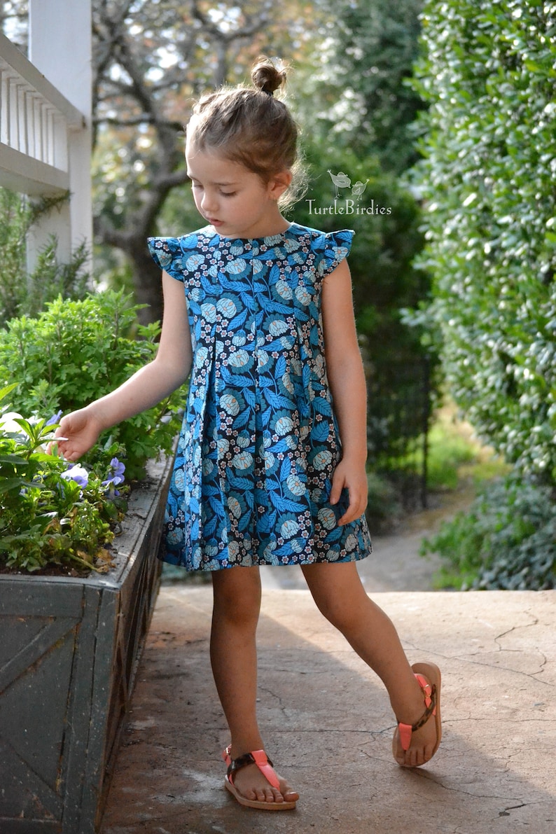 May include: A young girl wearing a blue and black floral dress with a ruffled neckline and short sleeves. She is standing on a sidewalk with a planter behind her.