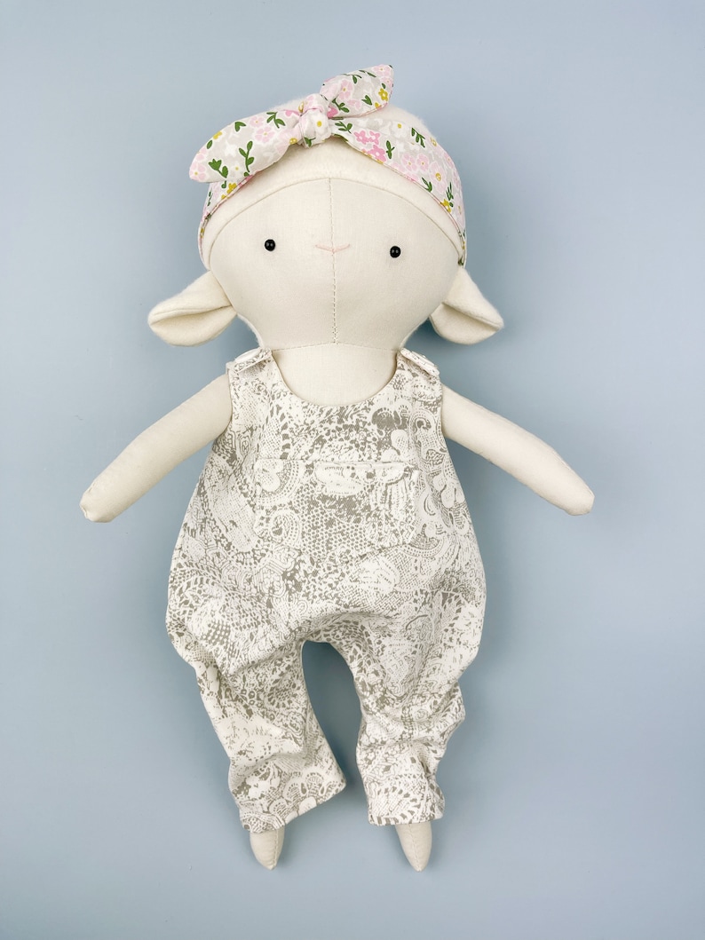 May include: A white plush lamb doll wearing a floral headband and a white and gray patterned jumpsuit.