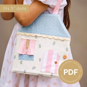 Könnte beinhalten: Stoff-Tragetasche in Form eines Puppenhauses mit blauem Blumendach und weißem Korpus. Die Tasche hat eine rosa und weiß gestreifte Tür, ein rosa Fenster mit Vorhängen und einen Holzknopfverschluss. Der Text "fits 5" dolls" ist sichtbar.