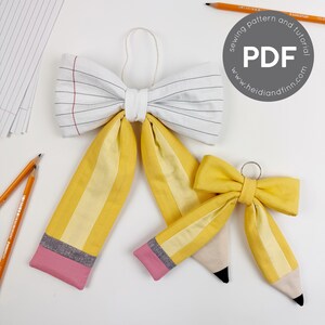 May include: Two fabric bows shaped like pencils and notebook paper. The larger bow features a white notebook paper pattern with black lines and a red margin, while both bows have yellow pencil bodies, grey metal bands, and pink eraser tips.