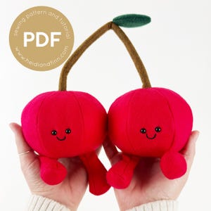 May include: Two red plush cherries with smiling faces and small legs. The cherries are connected by a brown stem with a green leaf.