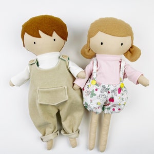 May include: Two handmade fabric dolls, one with brown hair wearing beige overalls and a white shirt, and the other with brown hair wearing a pink shirt and floral print shorts.