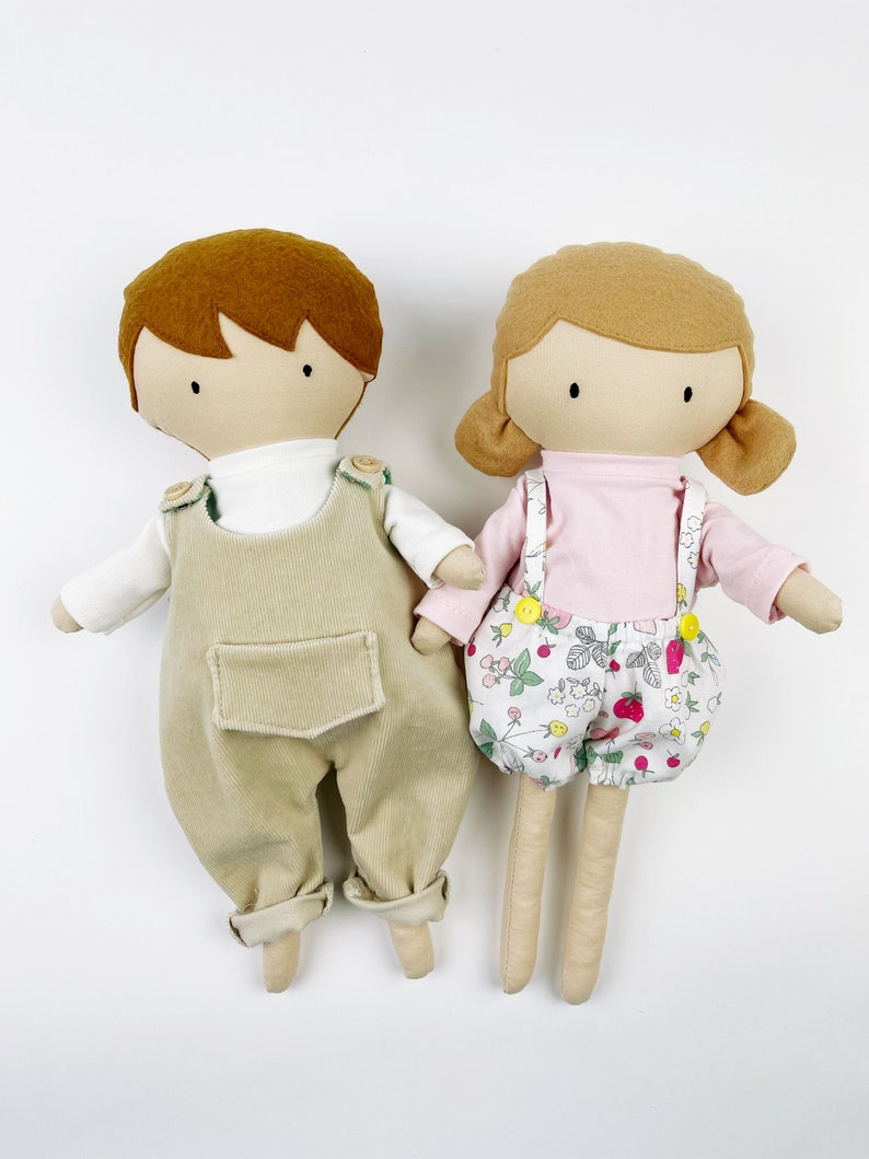 May include: Two handmade fabric dolls, one with brown hair wearing beige overalls and a white shirt, and the other with brown hair wearing a pink shirt and floral print shorts.
